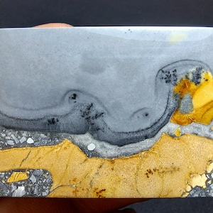 Puede incluir: Una losa de piedra rectangular con un patrón único. La parte superior es gris claro, pasando a un gris más oscuro con remolinos negros. La parte inferior presenta una mezcla de texturas doradas, grises y blancas, creando una apariencia de paisaje.