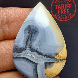May include: A pear-shaped gemstone displaying a distinctive pattern of grey, white, and yellow tones. The stone has a swirling design with dark grey and black lines. The text "US IMPORT TARIFF FREE GUARANTEE" is visible.
