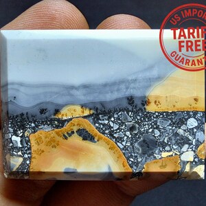May include: A rectangular gemstone with a landscape design. The top portion is light grey, transitioning to darker grey and black, with a yellow and brown section at the bottom. The stone has a polished surface and a natural, earthy appearance.