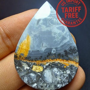May include: A teardrop-shaped gemstone with a unique pattern of grey, white, yellow, and black. The stone has a polished surface and a natural, layered appearance. The image includes a red circular stamp with the text "US IMPORT TARIFF FREE GUARANTEE".