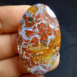 May include: An oval-shaped gemstone with a vibrant pattern of red, orange, yellow, white, and gray. The stone's surface displays a complex, natural design, resembling a landscape or abstract artwork. The polished gemstone is held in a hand.