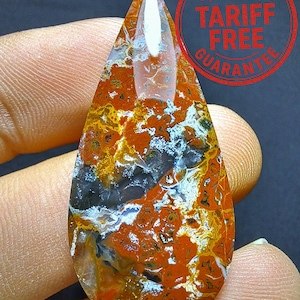 May include: A teardrop-shaped gemstone with a striking pattern of orange, yellow, black, and white. The stone is polished and translucent, with a pointed top. The image includes a red circular stamp with the text "US IMPORT TARIFF FREE GUARANTEE".