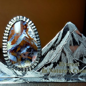 Pode incluir: Anel oval com moldura texturizada prateada. A pedra apresenta um design de paisagem com tons de castanho, branco e azul. O anel repousa sobre um suporte decorativo em forma de montanha com o texto "Everest In Nature's Creation".