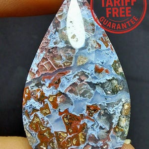 May include: A teardrop-shaped gemstone with a distinctive pattern of blue, red, gold, and white. The polished stone has a natural, organic appearance. The image includes a red circular stamp with the text "US IMPORT TARIFF FREE GUARANTEE".