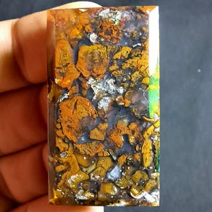 Può includere: Una gemma rettangolare con un mosaico di colori. La pietra presenta un mix di tonalità arancioni, marroni, grigie e bianche, con una qualità traslucida. La superficie ha un aspetto strutturato, con una finitura lucida. La gemma è tenuta in una mano.