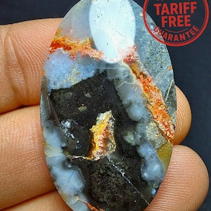 Può includere: Una gemma ovale con una miscela di colori, tra cui bianco, grigio, nero e arancione. La pietra ha una superficie lucida e un motivo unico. L'immagine include anche un cerchio rosso con il testo "US IMPORT TARIFF FREE GUARANTEE".