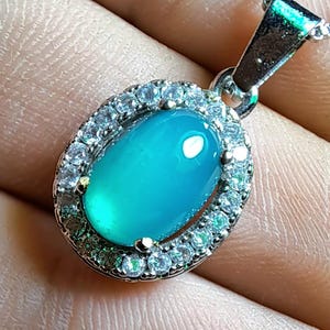 Puede incluir: Collar con colgante ovalado con una vibrante gema de color turquesa, rodeada por un halo de peque&ntilde;as gemas transparentes. El colgante est&aacute; suspendido de una delicada cadena de plata, creando una pieza de joyer&iacute;a cl&aacute;sica y elegante.