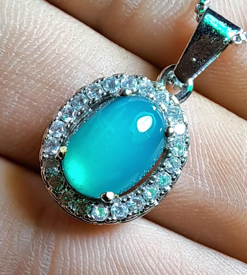 Puede incluir: Collar con colgante ovalado con una vibrante gema de color turquesa, rodeada por un halo de peque&ntilde;as gemas transparentes. El colgante est&aacute; suspendido de una delicada cadena de plata, creando una pieza de joyer&iacute;a cl&aacute;sica y elegante.