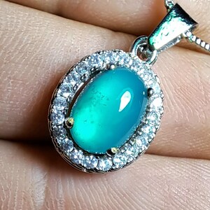 Puede incluir: Un collar con colgante de plata con una piedra preciosa ovalada de color turquesa. La piedra preciosa est&aacute; rodeada por un halo de peque&ntilde;os cristales transparentes. El colgante cuelga de una delicada cadena de plata, creando un look cl&aacute;sico y elegante.