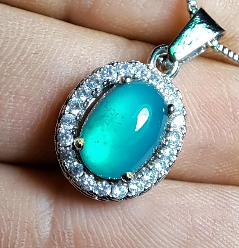 Puede incluir: Un collar con colgante de plata con una piedra preciosa ovalada de color turquesa. La piedra preciosa est&aacute; rodeada por un halo de peque&ntilde;os cristales transparentes. El colgante cuelga de una delicada cadena de plata, creando un look cl&aacute;sico y elegante.