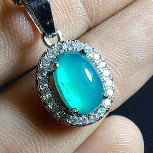 Puede incluir: Colgante de collar con una piedra preciosa ovalada de color turquesa. La piedra est&aacute; rodeada por un halo de peque&ntilde;as piedras redondas y transparentes engastadas en un metal plateado. El colgante cuelga de una cadena plateada.