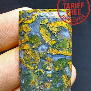 Op de afbeelding: Een rechthoekige edelsteen met een uniek patroon van goud, groen en blauw. De steen wordt in een hand gehouden, waarbij het gepolijste oppervlak wordt getoond. De afbeelding bevat een rode ronde stempel met de tekst "US IMPORT TARIFF FREE GUARANTEE".