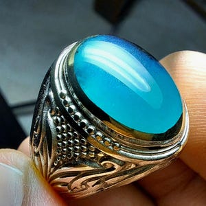 Peut inclure: Bague argentée ornée d'une grosse pierre ovale bleu clair. La pierre présente un effet dégradé, passant d'un bleu plus foncé en haut à une teinte plus claire. La monture de la bague est ornée de détails complexes.