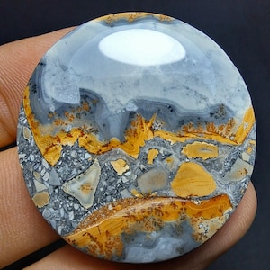 May include: A circular gemstone with a polished surface, displaying a distinctive pattern. The stone's design incorporates shades of grey, yellow, and brown, creating a marbled effect. The smooth surface reflects light, highlighting the natural colour and texture variations.