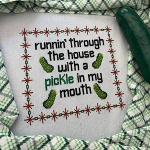 May include: Cross-stitch embroidery with the text "runnin' through the house with a pickle in my mouth" and pickle illustrations. A green pickle rests on a green and white patterned fabric.