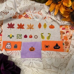May include: Cross-stitch sampler with autumnal and Halloween designs. The sampler includes leaves, pumpkins, and Halloween motifs like a ghost, spider, and witch's hat. The background is cream-coloured fabric, with a fall and Halloween theme.