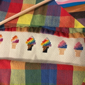 May include: A cross-stitch design featuring six ice cream cones with colorful scoops. The cones are stitched in various colors, including rainbow, pink, and purple. The design is set against a white background and framed by a rainbow plaid fabric.