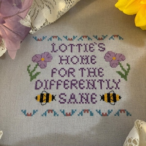May include: Cross-stitch embroidery with the text "Lottie's Home for the Differently Same." The design includes purple flowers, green stems, and two yellow and black bees. The background is a light fabric, with lace and flowers in the corners.
