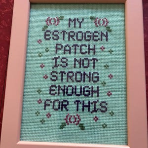 Może przedstawiać: Sztuka haftu krzyżykowego w jasnoróżowej ramie. Dzieło przedstawia tekst "MY ESTROGEN PATCH IS NOT STRONG ENOUGH FOR THIS" w ciemnofioletowej nici na jasnym zielonym tle, z kwiatowymi akcentami.