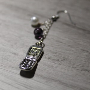 Può includere: Orecchino color argento con un ciondolo a forma di telefono a conchiglia. Il ciondolo del telefono è dettagliato con una tastiera e uno schermo. L'orecchino include anche una catena, una perla bianca e una perla viola scuro. Pende da un gancio d'argento.