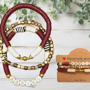 May include: Set of three bracelets with maroon, white, black, and gold beads. The bracelets are stacked on top of each other and spell out the word "AGGIES" in gold letters on white beads. The bracelets are on a brown card with the text "Handmade with Love" and "Best Wishes".