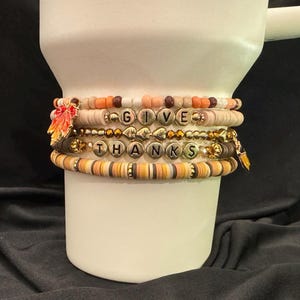 May include: Stack of beaded bracelets in fall colors, including orange, brown, and cream. The bracelets spell out "GIVE THANKS" with gold letter beads. Some bracelets have leaf charms.