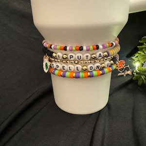 May include: A set of colorful beaded bracelets with the words "I PUT A SPELL ON U" in white letters. The bracelets are stacked on a white cup and include a black and green eye charm and a black cat charm.