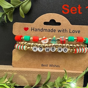 May include: Set of three Christmas-themed bracelets. The top bracelet has red, green, and gold beads with festive charms. The middle bracelet spells out "HOHOHO" with white letter beads. The bottom bracelet has red, green, and white striped beads.