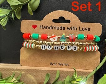 May include: Set of three Christmas-themed bracelets. The top bracelet has red, green, and gold beads with festive charms. The middle bracelet spells out "HOHOHO" with white letter beads. The bottom bracelet has red, green, and white striped beads.