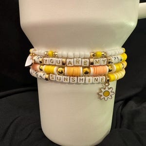 May include: A set of colorful beaded bracelets with the words "You are my sunshine" spelled out in white letters. The bracelets are stacked on top of each other and have a small daisy charm.