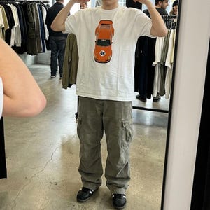 May include: White t-shirt featuring an orange race car graphic with the number 46. The shirt is paired with olive green cargo pants and black sneakers. The image is taken in a clothing store.