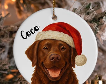 May include: A round, white Christmas ornament featuring a brown dog wearing a red Santa hat with a white trim. The dog's name, "Coco", is written in black script. A gold string is attached for hanging.