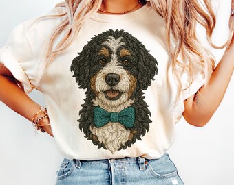 May include: A cream-colored t-shirt featuring a detailed graphic illustration of a Bernese Mountain Dog wearing a teal bow tie. The dog has black, brown, and white fur, brown eyes, and a friendly expression with its mouth slightly open.