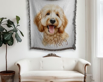 May include: A light gray throw blanket featuring a portrait of a golden doodle dog with its tongue out. The dog's name, "Riley", is written in a cursive font. The blanket has a dark gray fringe and is hanging above a white sofa.