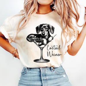 May include: A white t-shirt with a black and white illustration of a dachshund dog in a martini glass. The text "Cocktail Weiner" is printed on the shirt.