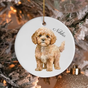 May include: A round white ceramic ornament featuring a detailed illustration of a light brown curly-haired puppy standing. The name Nikka is printed in black cursive script to the right of the puppy. A gold string is attached at the top.
