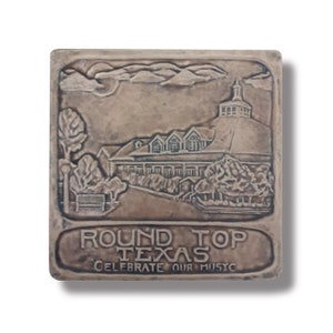 May include: A brown ceramic tile with a raised image of a building with a round roof and the text "ROUND TOP TEXAS CELEBRATE OUR MUSIC".
