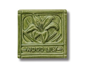 May include: A green ceramic tile with a raised design of a wood lily flower. The tile has a border and the words "WOOD LILY" are embossed at the bottom.