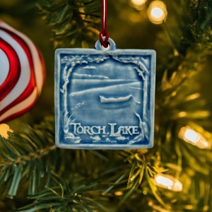May include: Blue and white ceramic Christmas ornament with a picture of a lake and a canoe. The ornament says "Torch Lake" on the bottom.