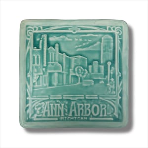 Könnte beinhalten: Eine hellblaue Keramikfliese mit einer Vintage-Illustration einer Stadtkulisse. Die Fliese hat einen dekorativen Rand und den Text "ANN ARBOR MICHIGAN" unten.