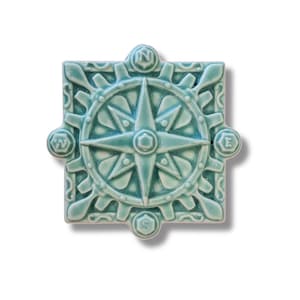 May include: A teal blue ceramic tile with a compass rose design. The compass rose has a star in the center and is surrounded by a circle of gears. The tile has the letters N, S, E, and W around the compass rose.