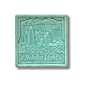 Puede incluir: Un azulejo de cerámica azul turquesa con una imagen en relieve blanca de un faro y una casa. El azulejo tiene el texto "Nobska Light CAPE COD" en relieve en la parte inferior.