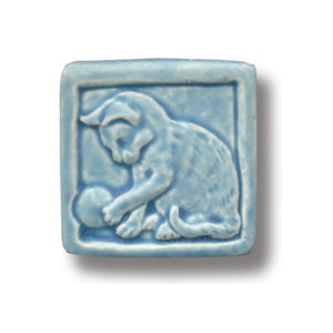 May include: A light blue ceramic tile featuring a raised image of a kitten playing with a ball. The tile is square shaped and has a slightly textured surface.