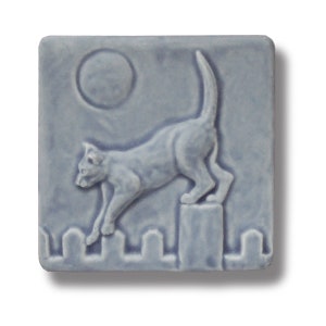 Puede incluir: Un azulejo de cerámica azul con una imagen en relieve de un gato saltando una valla. El gato está mirando hacia la izquierda y la luna es visible en el fondo.