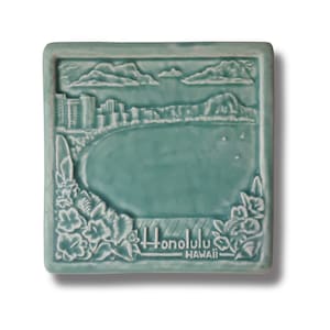 Puede incluir: Un azulejo de cerámica azul turquesa con una imagen en relieve de una ciudad y una masa de agua. El azulejo tiene un borde blanco y las palabras "Honolulu, Hawaii" están en relieve en la parte inferior.
