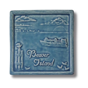 May include: A blue ceramic tile with a white raised image of a ferry boat, a seagull on a pier, and the words "Beaver Island".