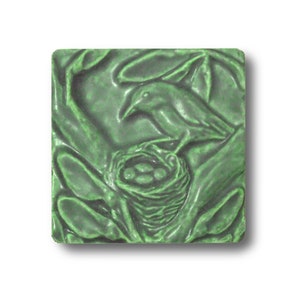 Puede incluir: Un azulejo de cerámica verde con un diseño en relieve de un pájaro sentado en un nido con huevos. El azulejo tiene una superficie texturizada y un aspecto rústico.
