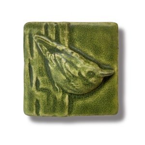 May include: A green ceramic tile with a raised image of a bird perched on a branch. The bird is facing the right side of the tile and has a detailed, textured design.