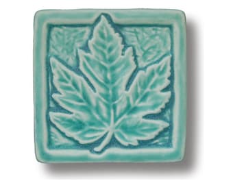 May include: A teal blue ceramic tile with a raised, embossed design of a single maple leaf.