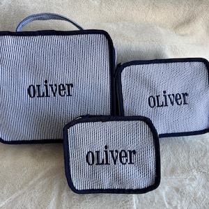 Puede incluir: Tres bolsas de aseo con cremallera de rayas azules y blancas con ribete negro. Las bolsas están personalizadas con el nombre "oliver" en bordado negro.